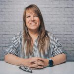 Cheerful female with crossed hands looking at camera while sitting at white table against brick wall